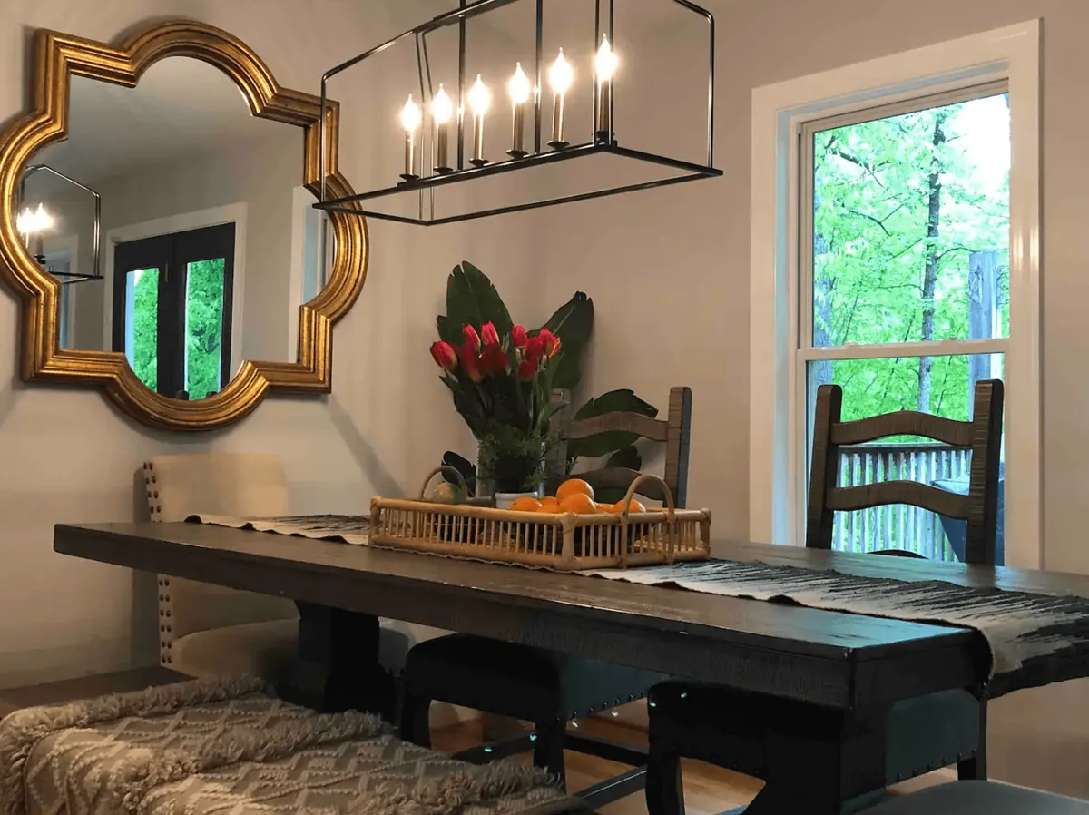 A dedicated dining area with a large table set under a statement chandelier, accented by a mirror and a fresh flower arrangement. The window provides natural light, making the space ideal for both casual breakfasts and special celebrations. The overall setup speaks to hospitality, conversation, and shared enjoyment.