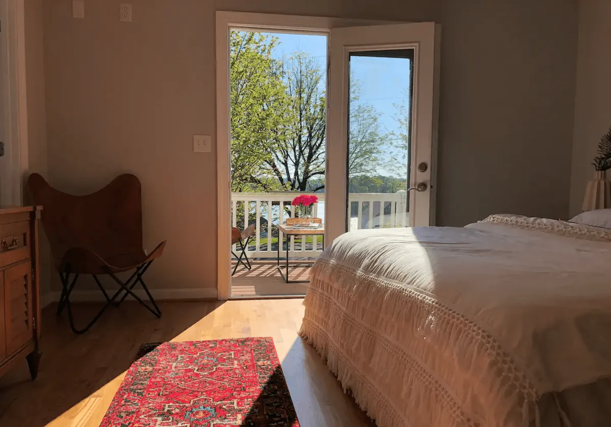 A cozy bedroom featuring a decorative blanket on the bed, a stylish butterfly chair, and open doors that lead directly onto a balcony. The room is filled with natural light, offering a peaceful retreat with inviting views of the outdoors.