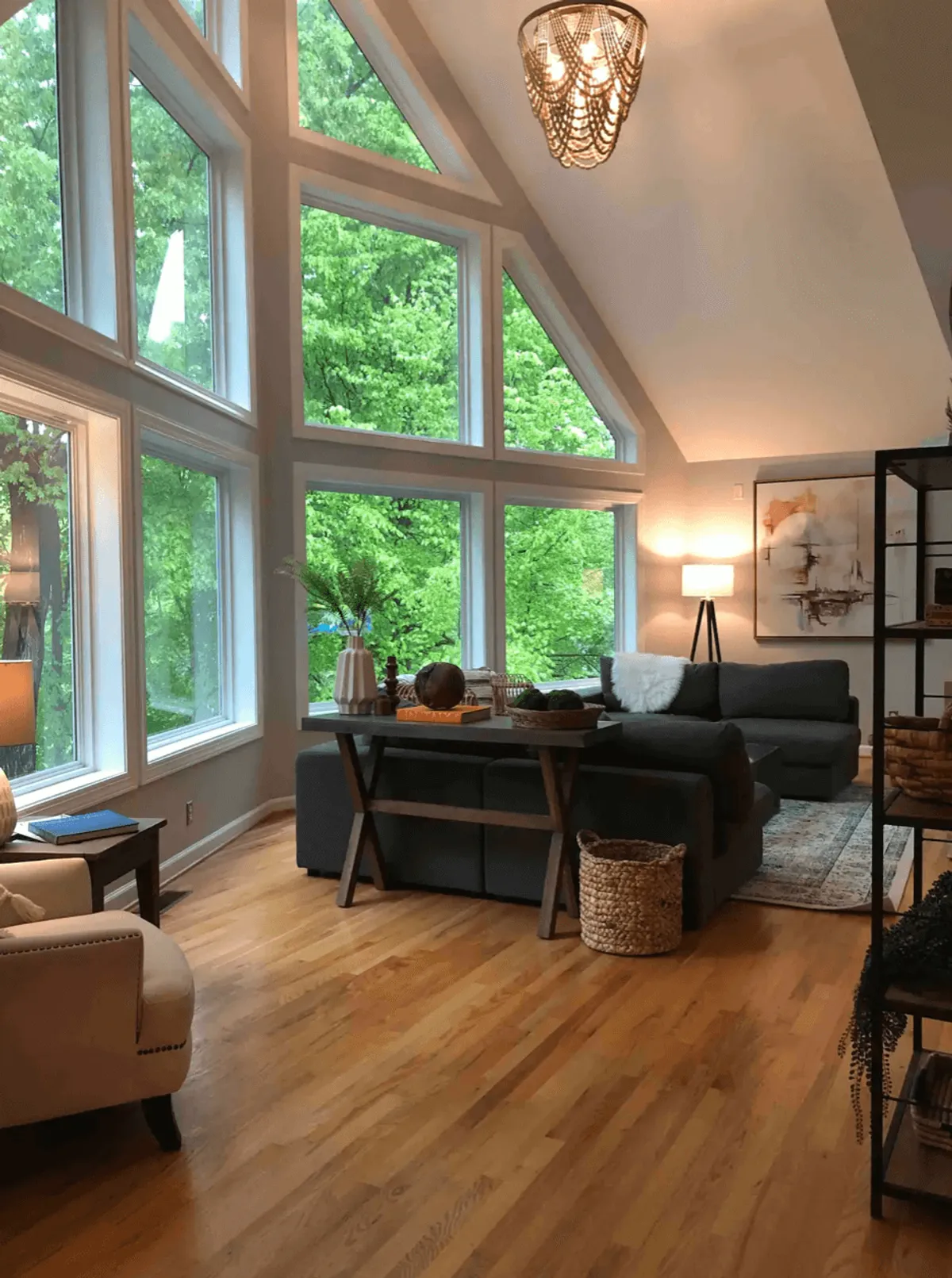 A cozy, inviting living space with a large sectional sofa, a wooden coffee table, and expansive windows overlooking lush greenery. Decorative vases and books on the table add warmth, while the view outside connects the indoors to nature.