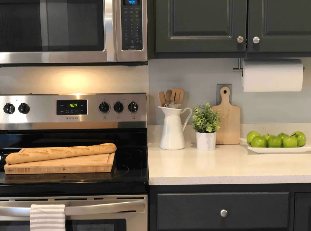 This image captures a warm, homey kitchen scene featuring an oven with a golden loaf of bread, a cluster of green apples, a white pitcher filled with utensils, and a small accent plant. The space exudes comfort and functionality, suggesting thoughtful details that enhance daily routines and relaxation.