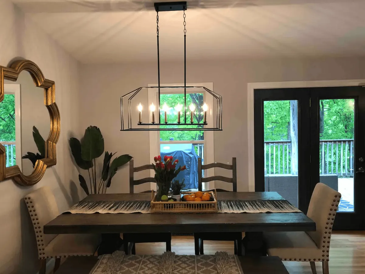 This interior image features an elegant dining area with a spacious table set beneath a glowing chandelier. Natural light streams in through a window, revealing a decorative mirror and a lush tropical plant nearby. The scene highlights a harmonious blend of style and comfort, perfect for shared meals or quiet moments.