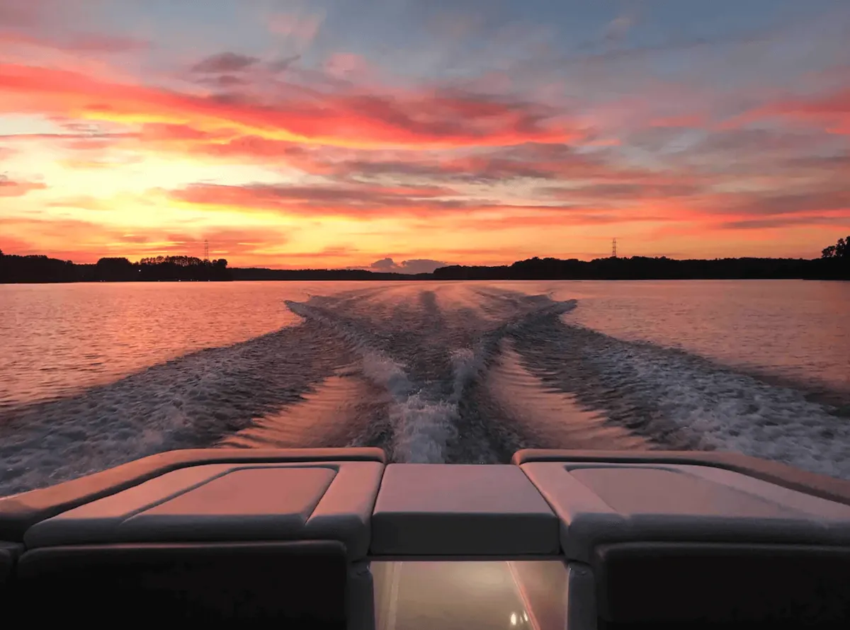 The hero image captures a vibrant Lake Norman sunset viewed from the back deck of a spacious lake house. Golden and pink hues reflect off tranquil waters, with cozy boat seating in the foreground, inviting guests to sit, relax, and savor the breathtaking view. The scene evokes a sense of peace and togetherness, perfect for unwinding or sharing memorable moments.